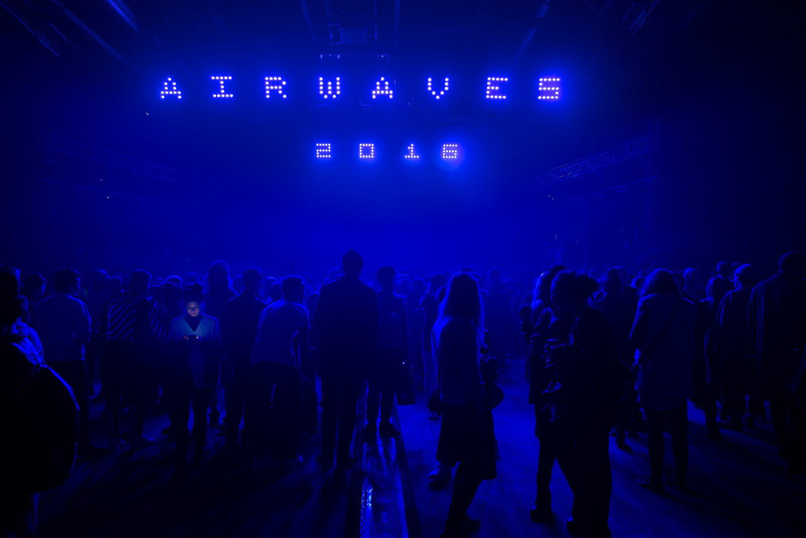 A photo of a crowd in a dark room illuminated by a bright blue neon sign saying "ICELAND AIRWAVES 2016"